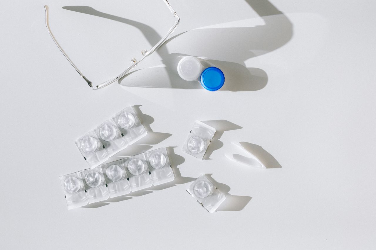 Flatlay of contact lens packages, case, and eyeglasses on a white surface with shadows.