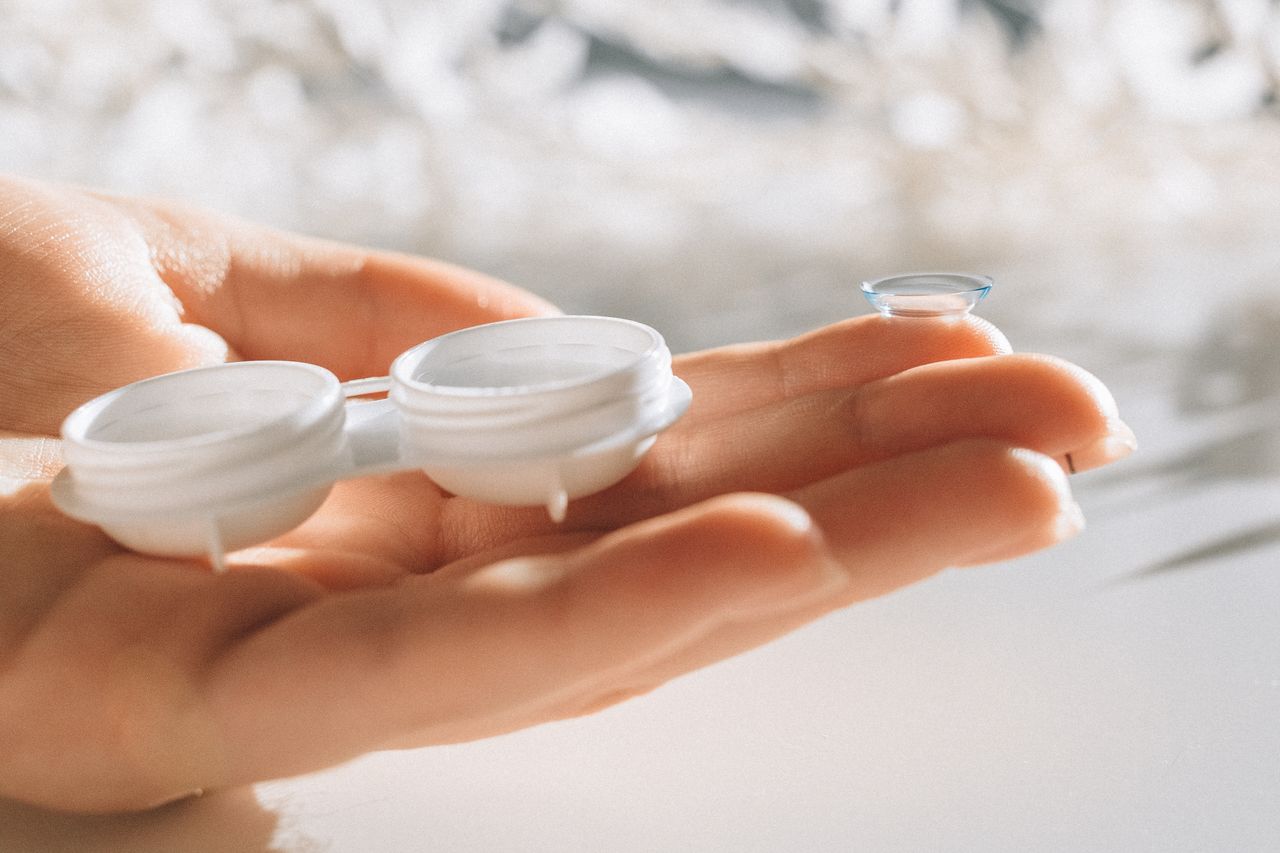 A hand delicately holds a contact lens and case against a blurred background.