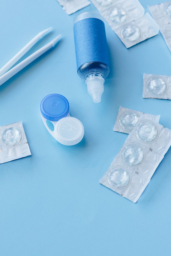 Flat lay image of a contact lens care kit including solution, lens container, and tweezers on a blue background.