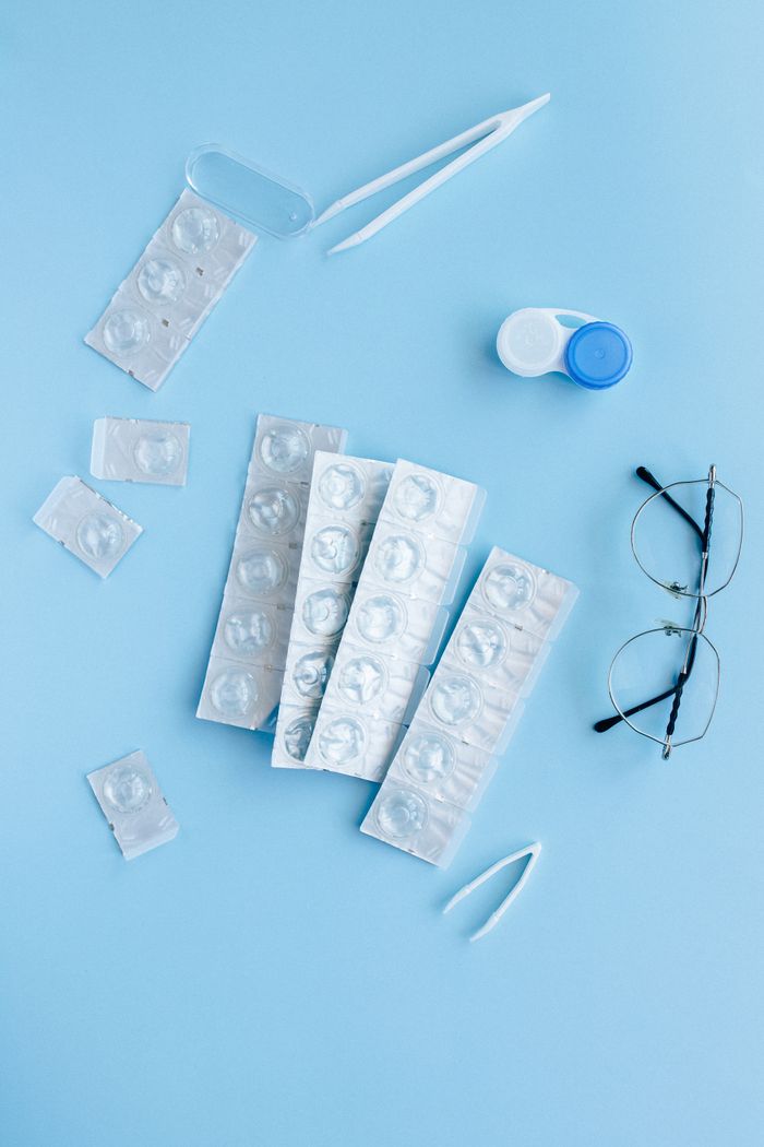 A top view flatlay of contact lenses, eyeglasses, and tweezers on a pastel blue background.