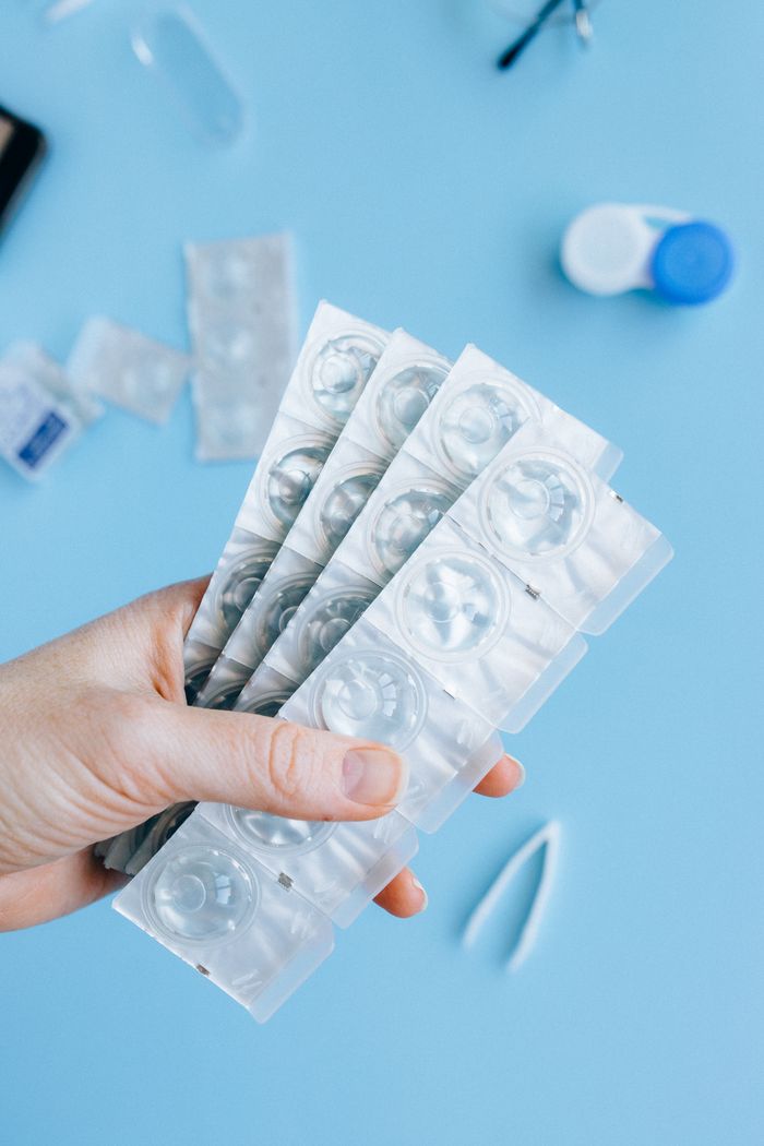 Hand holding blister packs of contact lenses with accessories on blue surface.